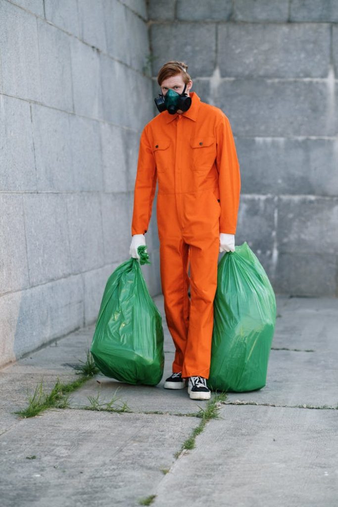 man-wearing-respirator-holding-green-plastic-bag-9246487 A man in an orange jumpsuit and mask carrying garbage bags against a concrete wall.